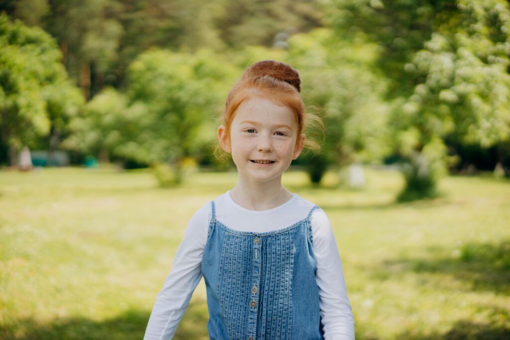 Une jeune fille rousse joyeuse sourit dans un parc ensoleillé et verdoyant, profitant d'une belle journée en plein air.