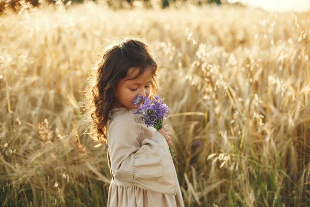 A young girl smells a purple flower bouquet in a vibrant wheat field during summer.